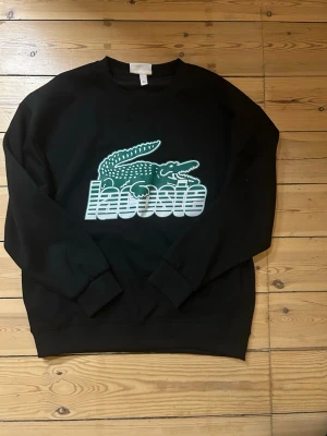Svart sweatshirt från Lacoste - Cool svart sweatshirt från Lacoste med stort grönt krokodillogo och vit text på bröstet. Tröjan har rund halsringning, långa ärmar och ribbade muddar. Perfekt för en avslappnad och trendig stil.