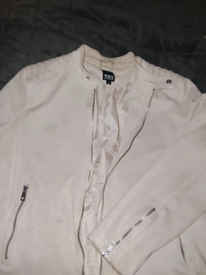 Beige skinnjacka från William Baxter - Snygg beige skinnjacka från William Baxter med raka dragkedjor, ståkrage med tryckknappar och detaljerade axlar. Jackan har dragkedjor vid ärmslut och fickor, samt en stilren och minimalistisk look. Perfekt för dig som gillar en clean och modern stil.