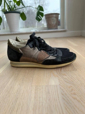 Svarta och bronsfärgade sneakers - Snygga sneakers i svart mocka och bronsfärgat skinn med snörning och beige sula. Hälpartiet har ett diskret leopardmönster som ger extra edge. Perfekta för dig som vill ha en unik och stilren look.