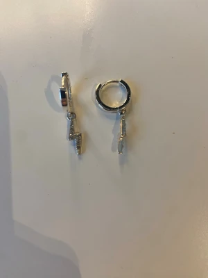 Silvriga blixtörhängen i hoops - Säljer ett par trendiga silvriga hoops med hängande blixtberlocker. Örhängena har en cool och modern design med små gnistrande stenar på blixtarna. Perfekta för dig som vill ha en edgy detalj till din look.