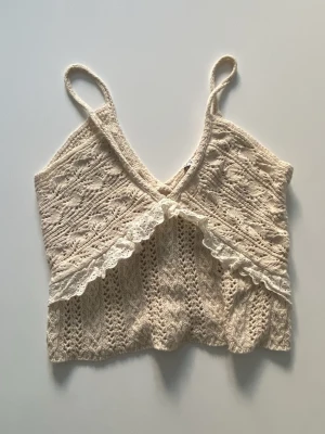 Beige virkad linnetopp med spets - Supersöt beige virkad linnetopp med smala axelband och omlott framtill. Toppen har fina spetsdetaljer längs kanten och är croppad i modellen. Perfekt för sommaren!