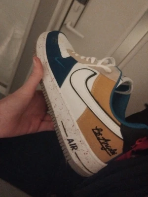 Nike Air Force 1 Los Angeles sneakers - Färgglada Nike Air Force 1 sneakers med vit, blå och gul ovandel, svart Swoosh och texten 'Los Angeles' på sidan. Skon har vit sula med röda stänk och klassisk snörning. Materialmix av skinn och syntet för en unik look.