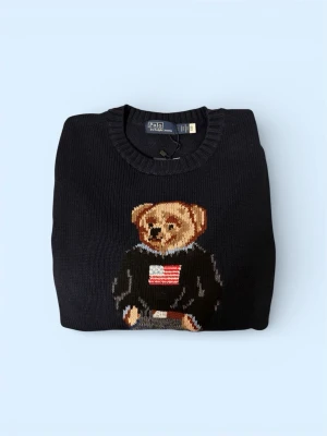 Svart stickad tröja Polo Bear - Svart stickad tröja från Polo Ralph Lauren med ikoniskt björnmotiv på bröstet. Tröjan har rund halsringning och ribbstickade muddar vid ärmslut och nederkant. Perfekt för dig som gillar klassiska och lekfulla detaljer.