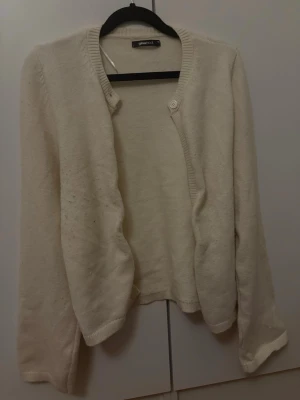 Beige kofta från Gina Tricot - Mysig beige kofta från Gina Tricot med rund halsringning och en knapp framtill. Koftan har långa ärmar och ribbade muddar vid ärmslut och nederkant. Perfekt att slänga över en topp för en enkel och stilren look. Behöver noppras lite.