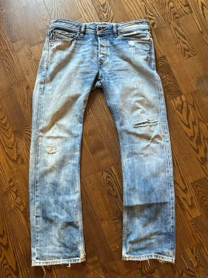 Blå Diesel Buster jeans med slitningar - Säljer ett par ljusblå Diesel Buster jeans med snygga slitningar och distressed detaljer. Klassisk femficksmodell med raka ben och låg midja. Jeansen är i bomull och har en avslappnad, casual vibe. Perfekta för dig som gillar en trendig och avslappnad stil.