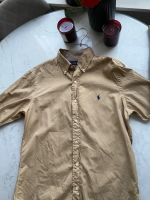 Beige skjorta från Ralph Lauren - Klassisk beige skjorta från Ralph Lauren med button-down krage och blå broderad logga på bröstet. Skjortan har långa ärmar och vita knappar framtill. Perfekt till en clean och stilren look. Nypris 1600kr. Ny utan prislapp, säljes då den var för liten.