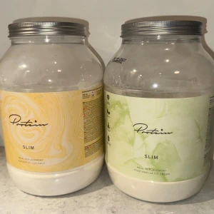 Protein pulver Slim måltidsersättning - Två stora burkar Protein Slim måltidsersättning i smakerna ananas/kokos och päron/vaniljglass. Burkarna är genomskinliga med färgglada etiketter, en gul och en grön, och har silverfärgade lock. Perfekt för dig som vill ha ett smidigt mellanmål.     Tagit 1 skopa från gul och 2 från grön. (Säljs pga allergi.) 