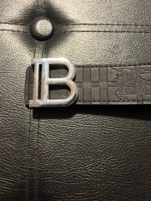 Svart Burberry bälte med B-spänne - Snyggt svart bälte från Burberry med ett stort silverfärgat B-spänne. Bältet har ett rutigt mönster och präglad Burberry-ryttare på lädret. Perfekt accessoar för att lyfta din outfit.