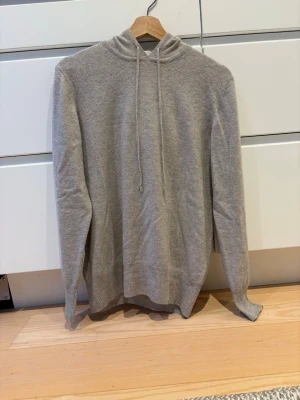 Soft Goat Kashmir hoodie - En stilren ljusgrå hoodie med dragsko i huvan och ribbade muddar vid ärmslut och nederkant. Hoodien har en enkel och clean look utan tryck eller logga, perfekt för en avslappnad stil. Materialet ser mjukt och bekvämt ut, troligen bomull eller bomullsblandning.