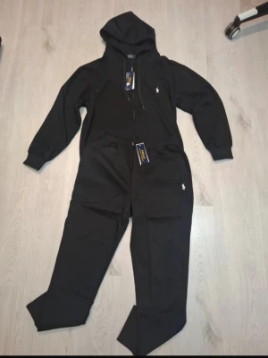 Svart jumpsuit med huva från Polo Ralph Lauren - Svart jumpsuit från Polo Ralph Lauren med huva och dragkedja framtill. Tillverkad i mjuk bomullsmix, har ribbade muddar vid ärmar och ben samt två fickor. Klassisk logga broderad på bröstet och benet. Perfekt för chill dagar eller när du vill vara extra bekväm.