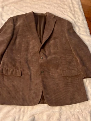 Brun manchesterkavaj med knappar - Snygg brun kavaj i manchester med klassisk krage och två knappar framtill. Kavajen har två fickor med lock och en bröstficka. Perfekt för dig som vill ha en retro och stilren look.