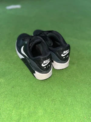 Nike Air Max 90 - Säljer ett par klassiska svarta Nike Air Max sneakers med vit sula och vita detaljer. Skorna har snörning och den ikoniska Nike-loggan på sidan och hälen. Perfekta för dig som gillar streetwear och sportig stil.