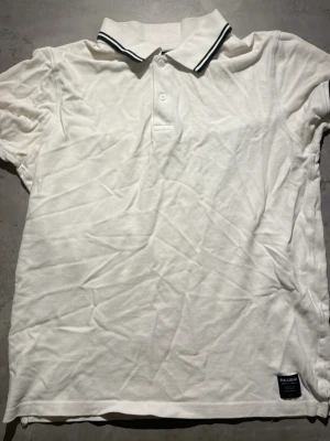 Vit pikétröja från Pull&Bear - Klassisk vit pikétröja från Pull&Bear med krage och två knappar framtill. Kragen har svarta ränder som detalj och tröjan är tillverkad i mjuk bomull. Perfekt till en avslappnad och stilren look.