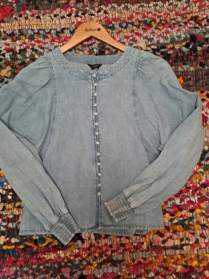 Ljusblå jeansblus från Lindex - Trendig ljusblå jeansblus från Lindex med långa ärmar och knappar framtill. Blusen har rund halsringning och smockade muddar vid ärmsluten. Tillverkad i mjukt denimtyg som ger en avslappnad men snygg look. Stl L men är som en M.