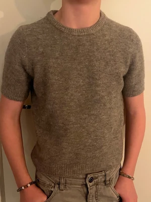 Grå stickad t-shirt i ullmix - Stickad t-shirt i beige ullmix med korta ärmar och rund hals. Tröjan har ribbstickade muddar vid ärmslut och nederkant för en snygg passform. Perfekt för lager-på-lager och en stilren look. Storlek S men passar Xs bättre. Pris kan diskuteras!🩷🫰🏻