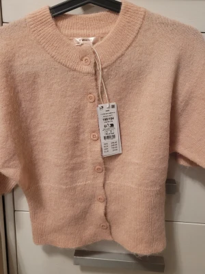 Beige kofta från Gina Tricot - Supersöt beige kofta från Gina Tricot med rund halsringning och korta ärmar. Koftan har en mjuk, stickad känsla och stängs med matchande knappar framtill. Perfekt för lager på lager och enkel att styla till olika outfits.