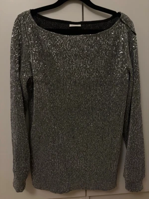 Glittrig silver topp med paljetter - Snygg långärmad topp i silver med massor av glittrande paljetter. Toppen har en rak passform och rund halsringning, perfekt för att sticka ut. Materialet känns mjukt och stretchigt mot huden.