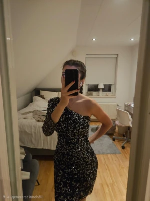 Svart glittrig one shoulder-klänning - Säljer en svart kortklänning med glittriga paljetter och one shoulder-design. Klänningen har en lång ärm på ena sidan och är ärmlös på den andra, vilket ger en unik och festlig look. Perfekt för dig som vill sticka ut.