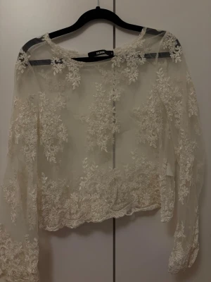 Vit spetsblus från Bik Bok - Superfin vit spetsblus från Bik Bok med transparent tyg och broderade blommor över hela plagget. Blusen har långa ärmar och en romantisk, bohemisk känsla. Perfekt att styla med jeans eller kjol för en trendig look.