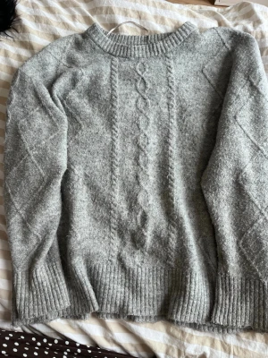Ljusgrå stickad tröja från 157 - Mysig ljusgrå stickad tröja från 157 med kabelstickat mönster framtill och ribbade muddar vid ärmslut och nederkant. Tröjan har rund halsringning och är perfekt för kyliga dagar. Klassisk och enkel design som passar till det mesta.