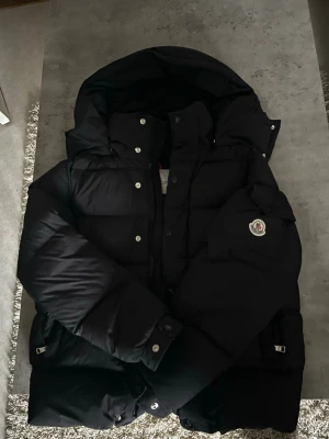 Moncler Vezere - Nyskick!! Snygg svart dunjacka från Moncler med huva och quiltad design. Jackan har långa ärmar, dragkedja och knappar framtill samt Moncler-logga på ärmen. Perfekt för kalla dagar och riktigt stilren look.