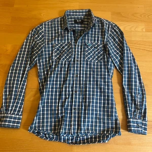 Blårutig skjorta från D&G - Snygg blårutig skjorta från D&G med klassisk krage och två bröstfickor med knappar. Skjortan har långa ärmar och är i tunn bomull, perfekt för dig som gillar en stilren men avslappnad look. Broderad D&G-logga på ena fickan ger extra detalj vet inte om den är riktig. 