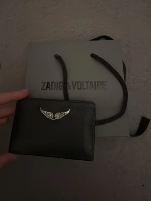 Svart plånbok från Zadig & Voltaire - Stilren svart plånbok från Zadig & Voltaire i skinn med silverfärgad vingdetalj på framsidan. Plånboken har flera kortfack på baksidan och en smidig, kompakt form som passar perfekt i fickan eller väskan.