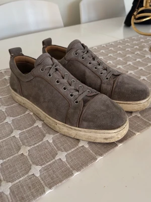 Grå mockasneakers med röd sula - Snygga grå sneakers i mocka från Christian Louboutin med klassisk röd sula. Skorna har rund tå, snörning och en stilren design som passar till det mesta. Perfekta för dig som gillar exklusiva detaljer och vill sticka ut med en ikonisk sula.