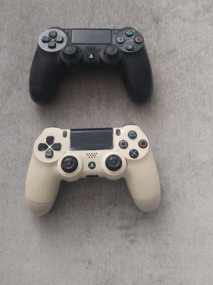 Svart och vit PlayStation DualShock 4 - Två PlayStation DualShock 4-handkontroller, en i svart och en i vit/beige. Klassisk design med touchpad, LED-ljus och ikoniska PlayStation-knappar. Perfekt för gaming på PlayStation-konsoler. Tillverkade i tålig plast med greppvänlig form.