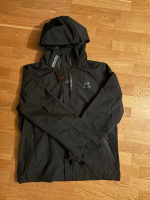 Svart vindjacka från Arc'teryx - Snygg svart vindjacka från Arc'teryx med huva och dragkedja framtill. Jackan har minimalistisk design, två sidofickor med dragkedja och logga på bröstet. Perfekt för blåsiga dagar och enkel att packa ner i väskan. Varan är ej äkta, storlek L men passar M bättre 