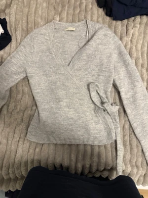 Ljusgrå omlottkofta från Gina Tricot - Säljer en ljusgrå stickad omlottkofta från Gina Tricot. Koftan har långa ärmar, ribbade muddar och knyts i sidan för en snygg passform. Storlek S och endast används ett fåtal gånger så känns som ny!!