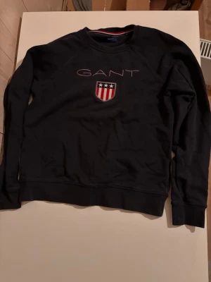 Svart sweatshirt från GANT - Snygg svart sweatshirt från GANT med broderad logga och sköld på bröstet. Tröjan har rund halsringning och långa ärmar. Skölden har röda och vita ränder samt tre vita stjärnor på blå botten. Perfekt till en avslappnad stil.