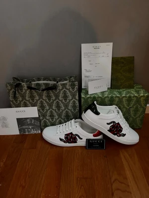 Gucci skor - Snygga vita sneakers från Gucci med ikoniskt orm-motiv i rött, svart och vitt på sidan. Skorna har snörning, rund tå och detaljer i grönt och rött på hälen. Tillverkade i skinn och levereras med originalkartong och dustbag. Pris kan diskuteras dom är i nyskick