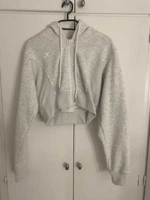Grå croppad hoodie med dragkedja - Snygg ljusgrå croppad hoodie med huva och half zip-dragkedja framtill. Hoodien har långa ärmar, ribbade muddar och snörning i huvan. Perfekt för en chill och sportig look.