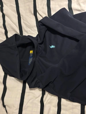 Mörkblå hoodie från Polo Ralph Lauren - Snygg mörkblå hoodie från Polo Ralph Lauren med klassisk huva och den ikoniska ljusblå polospelaren broderad på bröstet. Tillverkad i mjukt material som känns skönt mot huden. Perfekt för en avslappnad och stilren look.