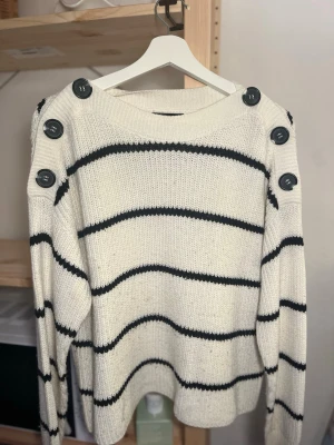 Vit stickad tröja med svarta ränder - Vit stickad tröja med svarta horisontella ränder och stora svarta knappar längs axlarna. Tröjan har rund halsringning och långa ärmar. Perfekt för en avslappnad och trendig look.