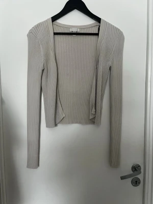 Beige ribbad öppen kofta - Säljer en beige, ribbad och tunn öppen kofta med knappar och lång ärm. Modellen är croppad och har en smal passform som sitter snyggt på kroppen. Perfekt att slänga över ett linne eller topp för en enkel men stilren look. Den är i bra skick och passar flera storlekar.