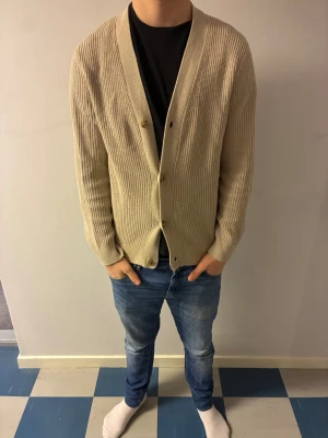 Kofta East West - Säljer en beige ribbstickad kofta från East West med v-ringning och tre knappar framtill. Koftan har lång ärm och en avslappnad passform, perfekt att slänga över en t-shirt. Personen på bilden är 183 hör av er vid frågor.