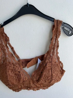 Brun spets-bh storlek M från Kappahl  - Snygg brun bh i spets med blommigt mönster och breda axelband. Bh:n har justerbara band och hakknäppning bak. Materialet är genombruten spets framtill och slät tyg på insidan av kuporna. Perfekt för dig som gillar en feminin och trendig look.