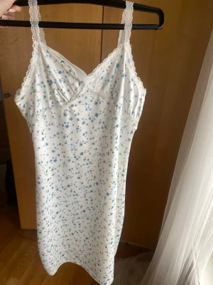 Blommig vit klänning med spets - Sött vitt klänning med tunna axelband och spetsdetaljer vid urringningen. Tyg med små blå blommor över hela plagget ger en romantisk känsla. Mjuk och lätt kvalitet, perfekt för mysiga kvällar hemma.