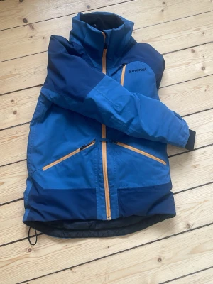 Blå vindjacka från Everest - Snygg blå vinterjacka från Everest med orange dragkedjor och hög krage. Storlek 146. Jackan har två snedställda fickor framtill och är tillverkad i slitstark polyester. Perfekt för blåsiga dagar och outdoor-aktiviteter.