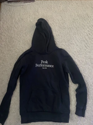 Mörkblå hoodie från Peak Performance - Snygg mörkblå hoodie från Peak Performance med vit logga på bröstet. Perfekt för en avslappnad och sportig stil. Mjuk bomullsblandning som känns skön mot huden.
