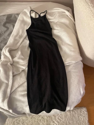 Svart ribbad långklänning - Snygg svart långklänning med smala axelband och ribbad struktur. Klänningen har en kroppsnära passform och är perfekt för dig som gillar en stilren och enkel look. Tillverkad i mjukt material som sitter skönt mot kroppen.