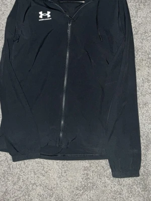 Svart vindjacka från Under Armour - Snygg svart vindjacka från Under Armour med dragkedja framtill och logga på bröstet. Jackan har lång ärm och är tillverkad i lätt polyester, perfekt för blåsiga dagar. Enkel och sportig design som passar till det mesta.