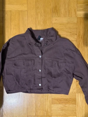 Croppad brun jeansjacka - Snygg croppad jeansjacka i mörkbrun färg från H&M. Jackan har klassisk krage, två stora bröstfickor med lock och knäppning framtill med silvriga knappar. Perfekt för en trendig och avslappnad look.