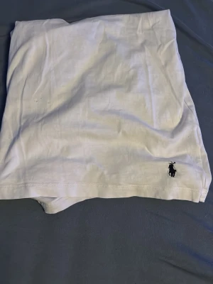 Vit t-shirt från Polo Ralph Lauren - Klassisk vit t-shirt från Polo Ralph Lauren med rund hals och liten broderad logga i svart vid nederkanten. Tillverkad i mjuk bomull som känns skön mot huden. Perfekt basplagg för en clean och stilren look.