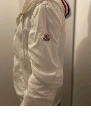 Vit Moncler vindjacka med huva - Snygg vit vindjacka från Moncler med huva och dragkedja framtill. Jackan har Moncler-logga på ärmen, snörning vid huvan och praktiska fickor med knappar. Perfekt för dig som vill ha en stilren och sportig look.