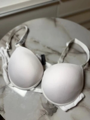 Vit BH med strassband från Victoria's Secret - Säljer en vit BH från Victoria's Secret med vadderade kupor och justerbara axelband dekorerade med strass och texten 'Victoria's Secret'. Snygg och stilren design som ger en exklusiv känsla.