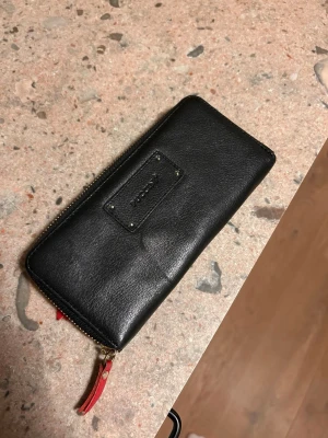 Svart skinnplånbok från Puccini - Stilren svart plånbok från Puccini i äkta skinn. Plånboken har guldfärgad dragkedja och en röd detalj på dragfliken. Rektangulär form med fack för både kort (totalt 12 st) och sedlar. Perfekt för dig som vill ha både stil och funktion. Mått: 20x8,5x2 cm. 