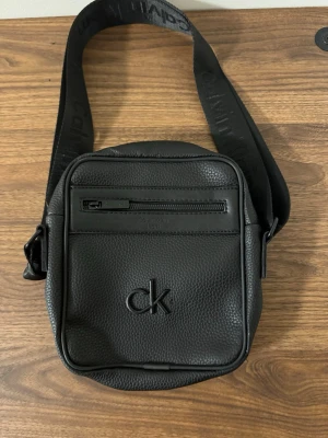 Svart axelväska från Calvin Klein - Snygg svart axelväska från Calvin Klein i strukturerat skinn. Väskan har justerbar axelrem med logga, dragkedja fram och bak samt diskret CK-logo på framsidan. Perfekt för att bära det viktigaste med stil.